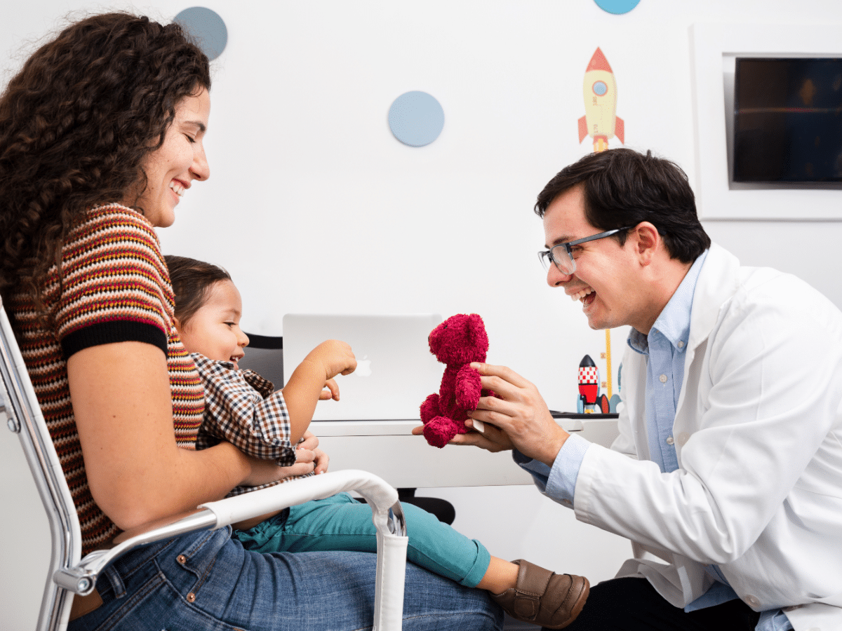  After providing flu treatment, a pediatrician in Fresno gives a teddy bear to his patient, a child sitting on their mother’s lap.
