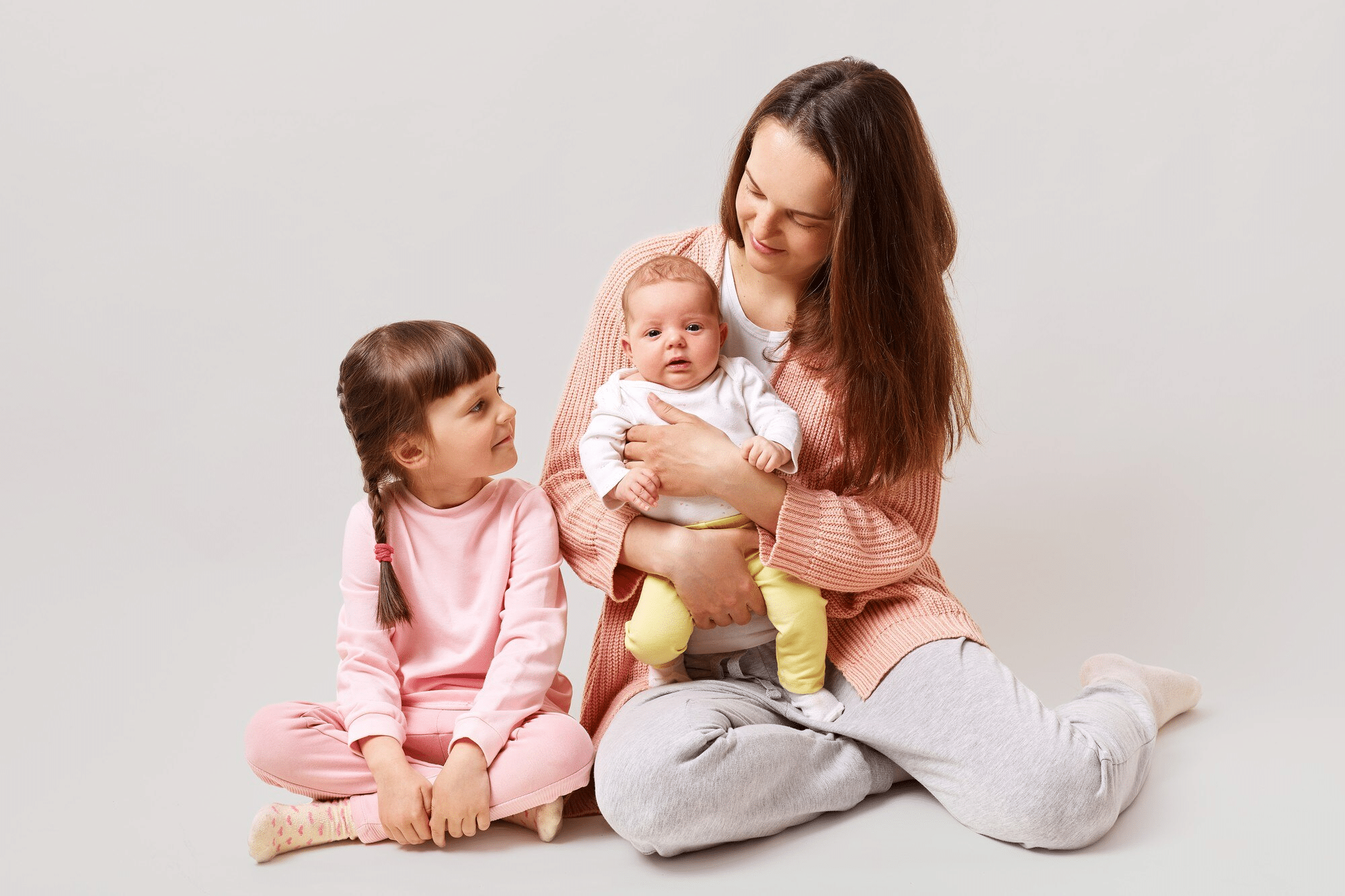 A mother holds her breastfeeding infant as her toddler sits beside her. 