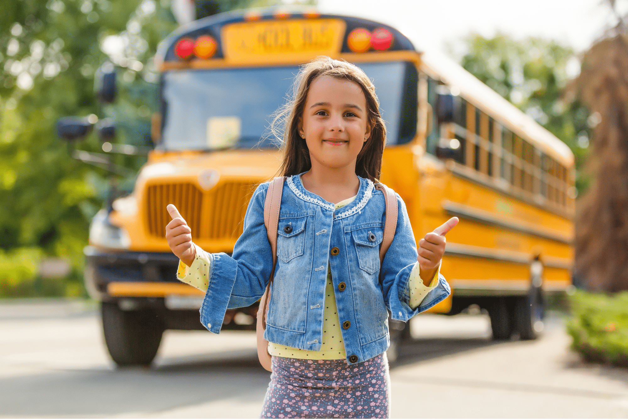 Little girl posing in front of a school bus on her first day after completing her back-to-school physical exam.